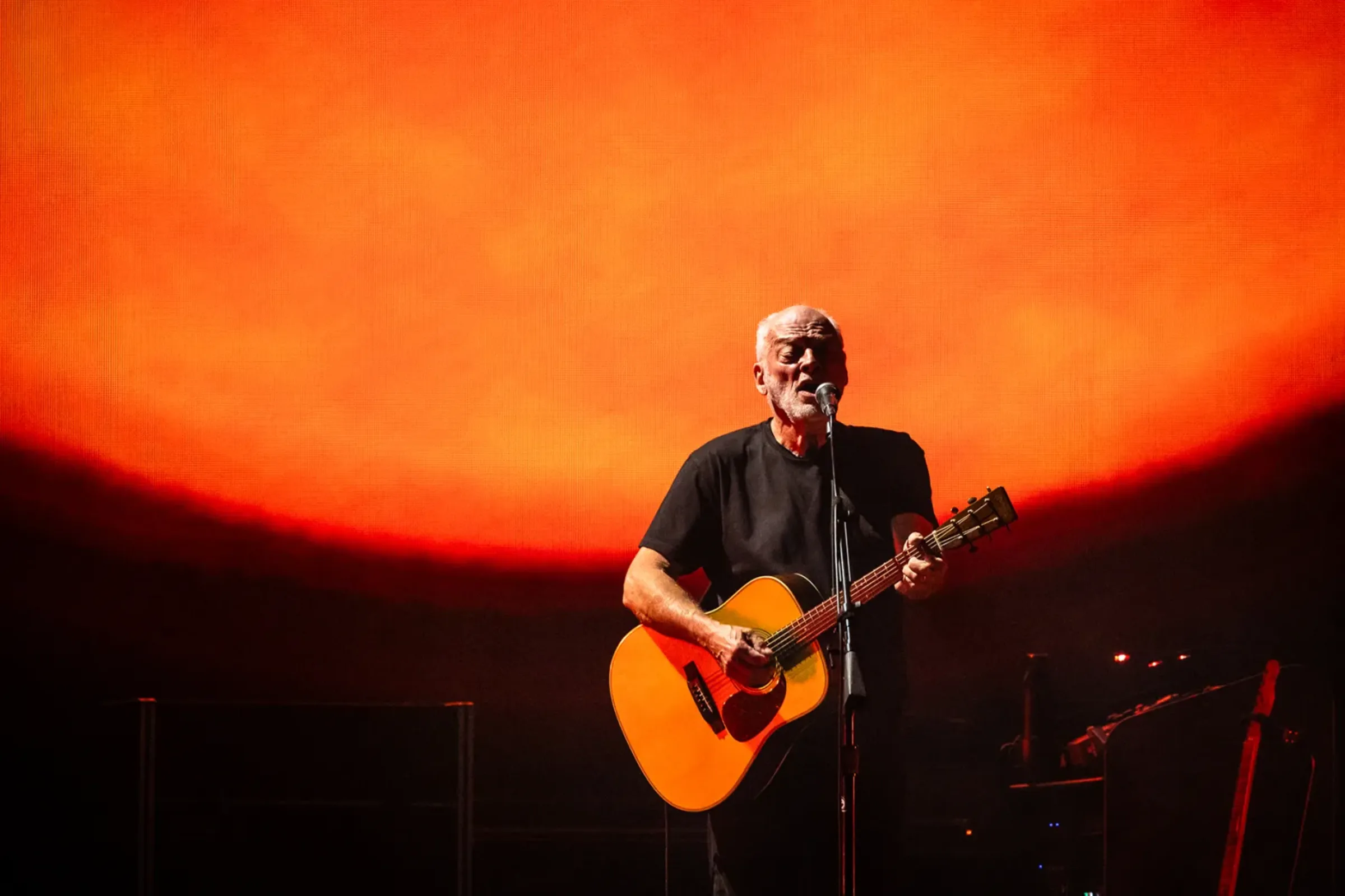 David Gilmour performing with acoustic guitar against an orange backdrop